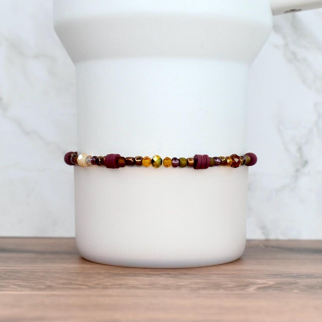 A beaded bracelet with a mix of burgundy, gold, and olive green beads. The bracelet features a variety of shapes and sizes, including small round beads and larger cylindrical beads.