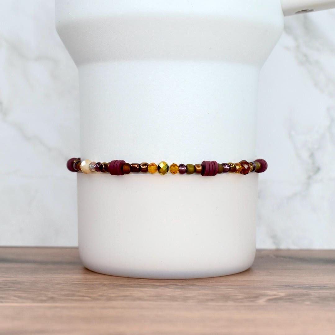 A beaded bracelet with a mix of burgundy, gold, and olive green beads. The bracelet features a variety of shapes and sizes, including small round beads and larger cylindrical beads.
