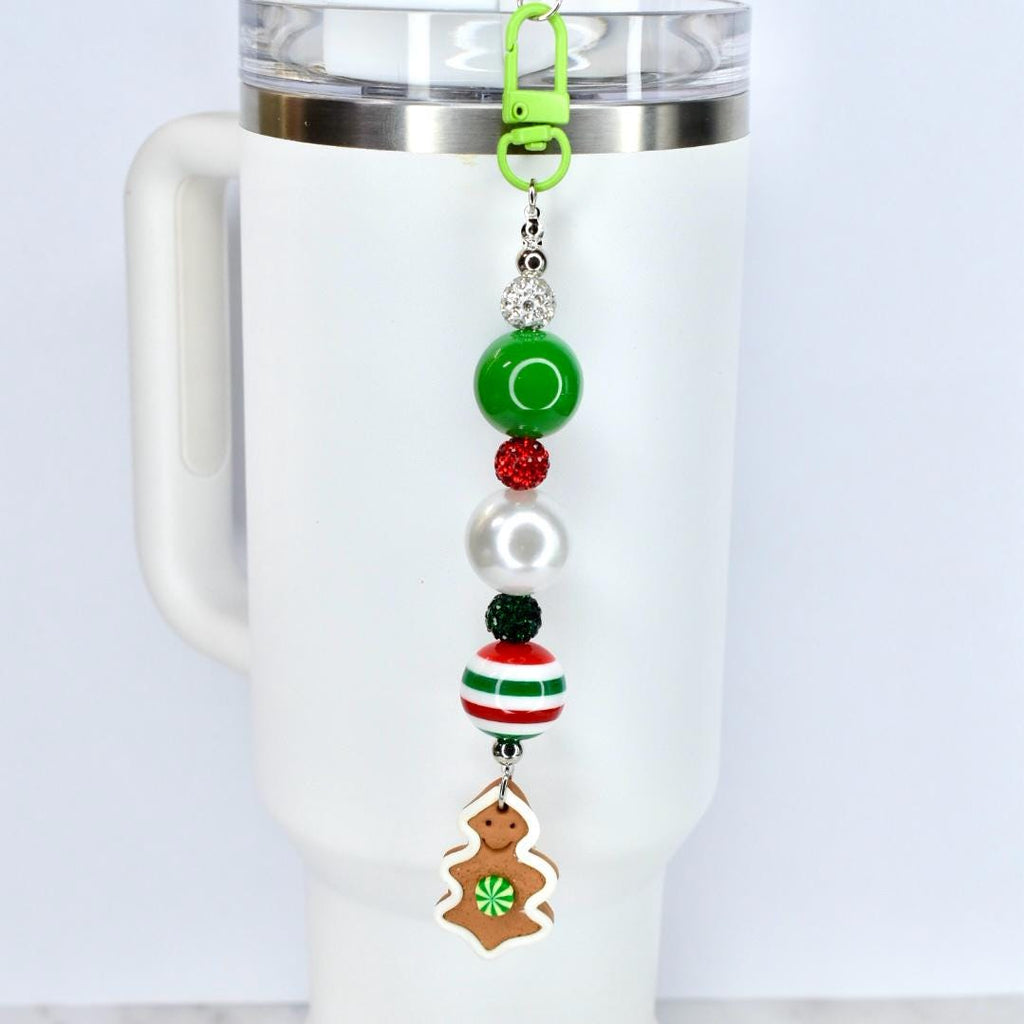 A beaded charm with a green clasp, attached to a white tumbler. The charm features a series of beads in green, red, white, and black, culminating in a gingerbread man-shaped charm with a green flower.