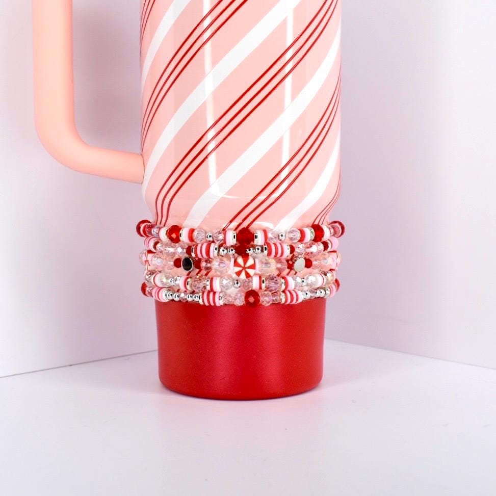 A red and white striped Stanley tumbler with a pink handle stands on a white surface. A set of five beaded bracelets with red, white, and clear beads encircles the base, adding a festive touch.