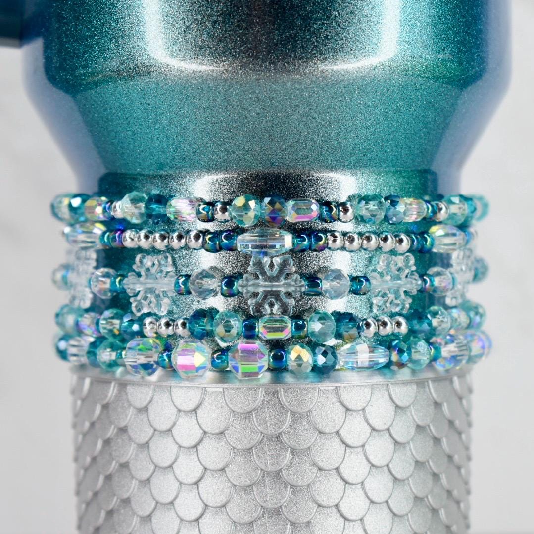 A teal tumbler with a silver, scaly base and a beaded band. The band features clear and iridescent beads, silver accents, and snowflake-shaped embellishments. The top of the tumbler has a glittery finish.
