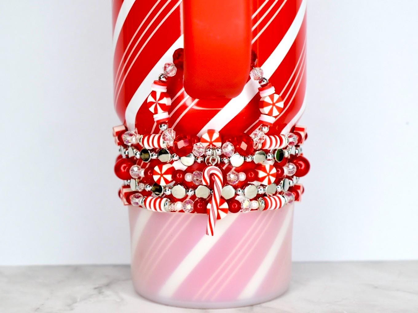 A red and white striped mug with a stack of red, white, and silver beaded bracelets. The bracelets feature candy cane charms and snowflake-shaped beads. The mug has a red handle and a pink and white striped base.