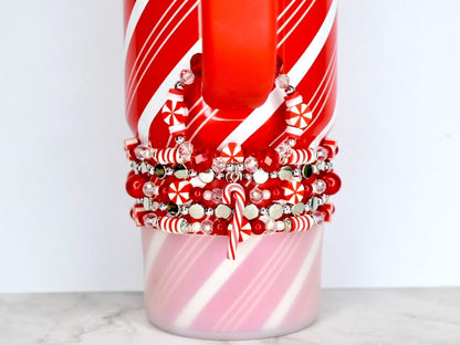 A red and white striped mug with a stack of red, white, and silver beaded bracelets. The bracelets feature candy cane charms and snowflake-shaped beads. The mug has a red handle and a pink and white striped base.