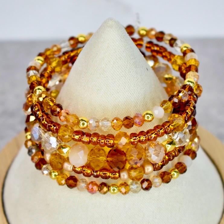 A stack of beaded bracelets in shades of gold, amber, and clear. The bracelets are displayed on a white cone-shaped stand. The beads vary in size and shape, with some featuring a faceted design.
