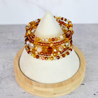 A stack of beaded bracelets in shades of amber, gold, and clear crystals. The bracelets are displayed on a white cone-shaped stand atop a wooden base. The beads vary in size and shape, creating a layered effect.