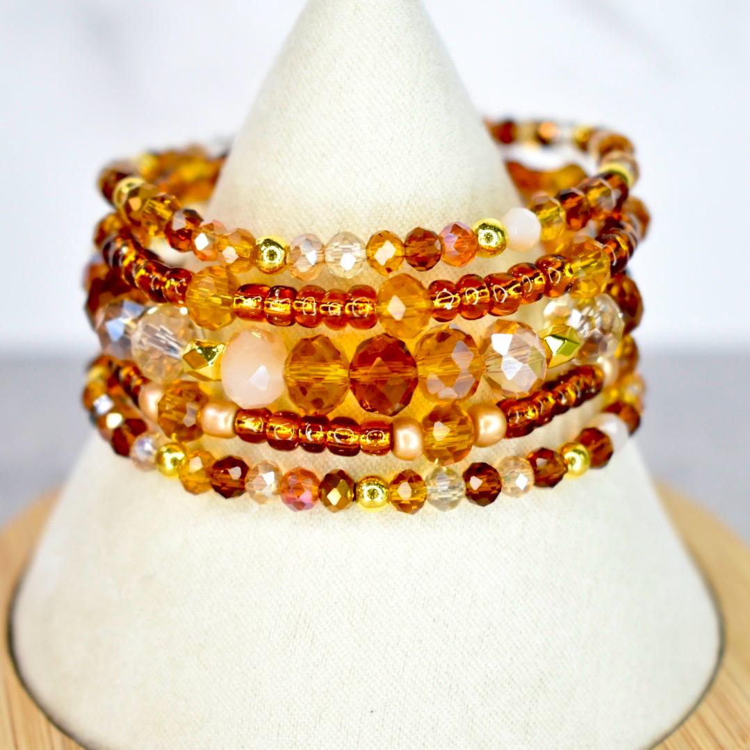 A stack of beaded bracelets in shades of amber, gold, and brown. The bracelets feature a variety of bead sizes and shapes, including small round beads and larger faceted beads. The bracelets are displayed on a white cone-shaped stand.
