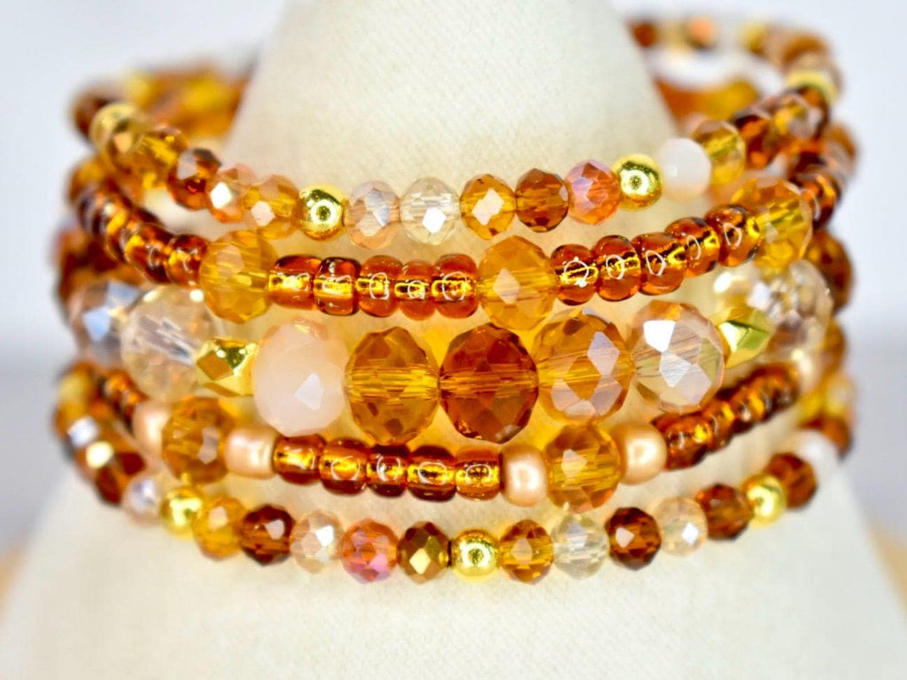 A stack of beaded bracelets in shades of amber, gold, and peach. The bracelets feature a variety of bead sizes and shapes, including small seed beads, faceted crystals, and round beads. The bracelets are displayed on a white surface.