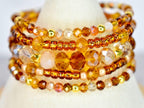 A stack of beaded bracelets in shades of amber, gold, and peach. The bracelets feature a variety of bead sizes and shapes, including small seed beads, faceted crystals, and round beads. The bracelets are displayed on a white surface.