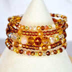 A stack of beaded bracelets in shades of amber, gold, and brown. The bracelets feature a variety of bead sizes and shapes, including faceted crystals, small round beads, and larger, opaque beads. The bracelets are displayed on a white cone.