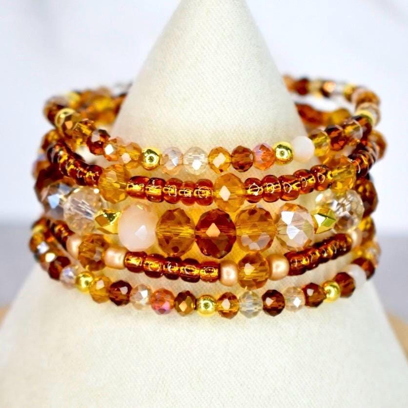 A stack of beaded bracelets in shades of amber, gold, and brown. The bracelets feature a variety of bead sizes and shapes, including faceted crystals, small round beads, and larger, opaque beads. The bracelets are displayed on a white cone.