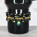 A black beaded bracelet set with gold and iridescent beads. The bracelets are stacked on top of each other and feature a teardrop-shaped green and blue crystal pendant.