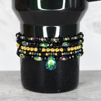 A black beaded bracelet set with gold and iridescent beads. The bracelets are stacked on top of each other and feature a teardrop-shaped green and blue crystal pendant.