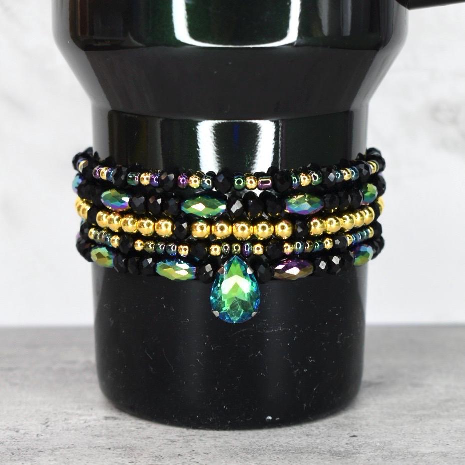 A black beaded bracelet set with gold and iridescent beads. The bracelets are stacked on top of each other and feature a teardrop-shaped green and blue crystal pendant.