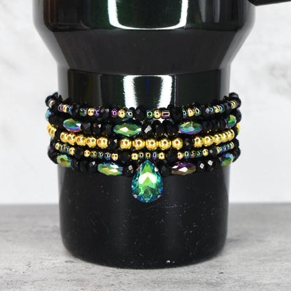 A black beaded bracelet set with gold and iridescent beads. The bracelets are stacked on top of each other and feature a teardrop-shaped green and blue crystal pendant.