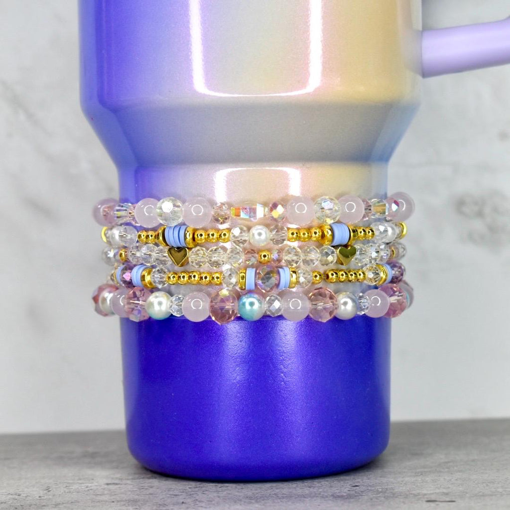 A stack of beaded bracelets in shades of pink, clear, and gold, with small heart and disc accents. The bracelets are wrapped around a purple and iridescent tumbler. The tumbler has a handle and a gradient finish.