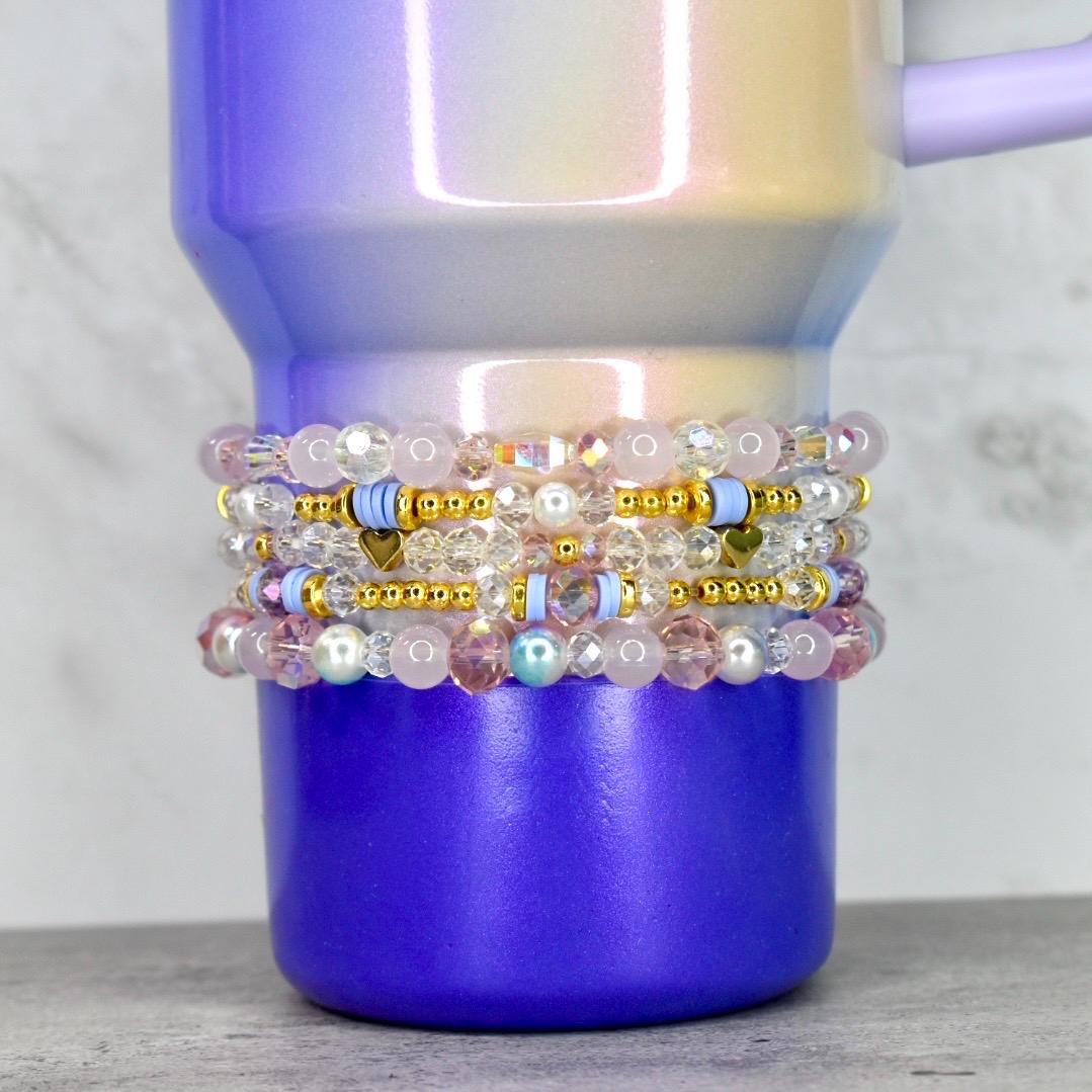 A stack of beaded bracelets in shades of pink, clear, and gold, with small heart and disc accents. The bracelets are wrapped around a purple and iridescent tumbler. The tumbler has a handle and a gradient finish.