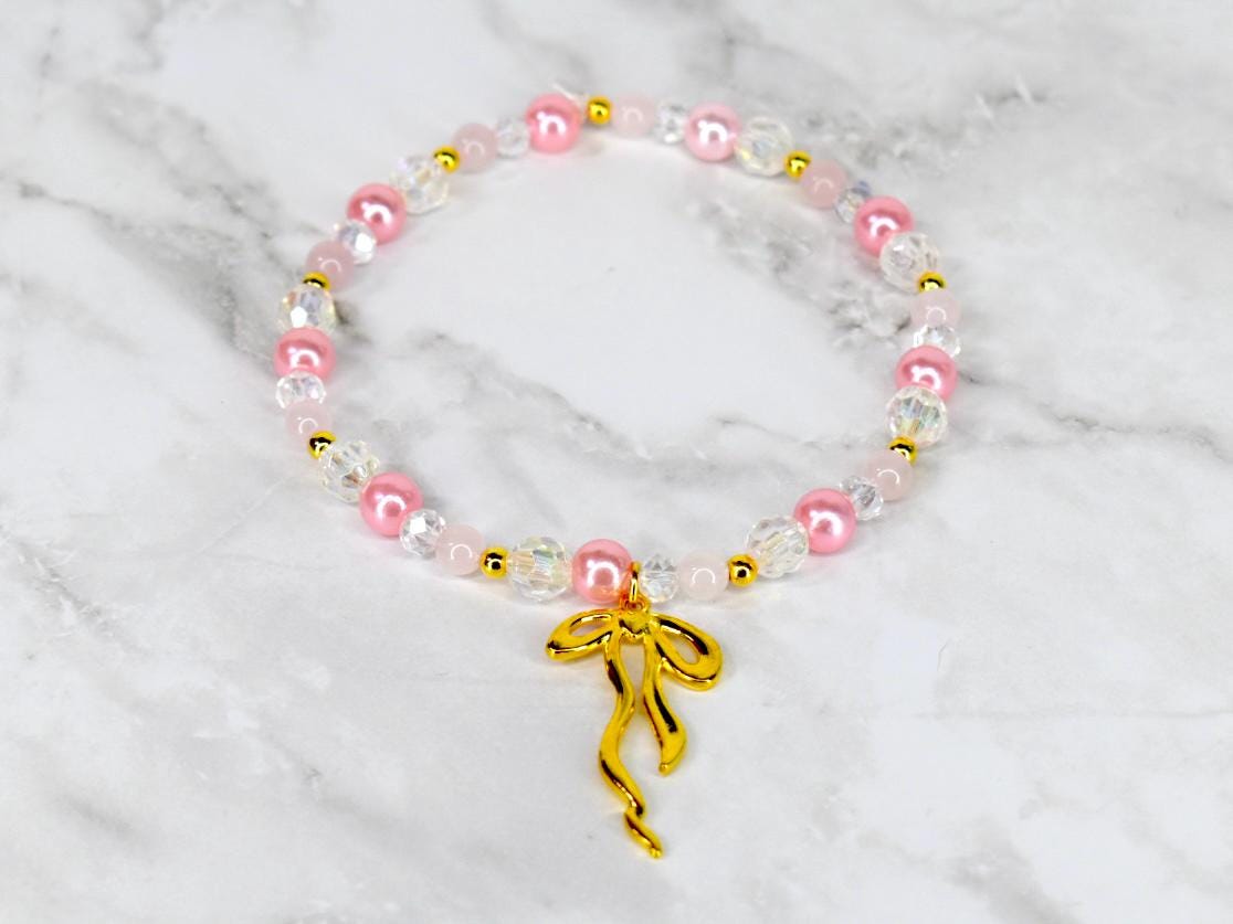 A beaded bracelet featuring pink, clear, and gold beads. The bracelet has a gold bow charm. The beads are strung together with a clear elastic cord. The bracelet is displayed on a white and gray marble surface.