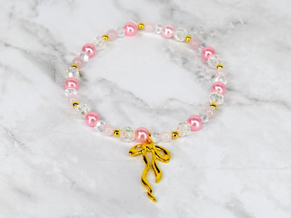 A beaded bracelet featuring pink, clear, and gold beads. The bracelet has a gold bow charm. The beads are strung together with a clear elastic cord. The bracelet is displayed on a white and gray marble surface.