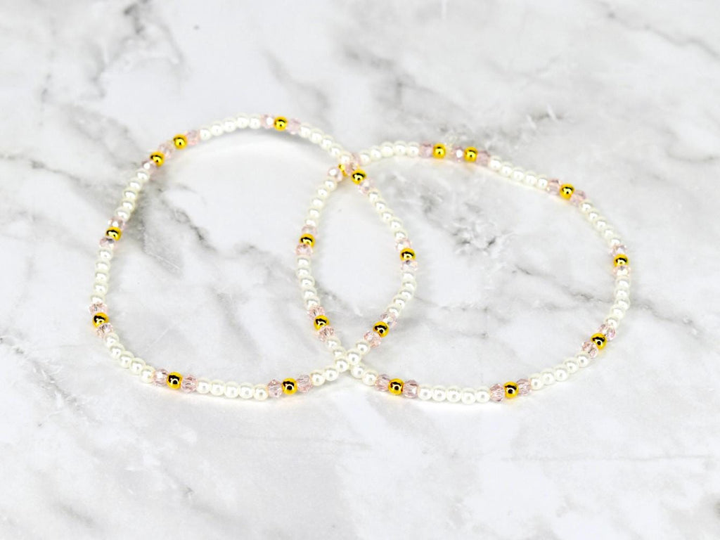 Two delicate beaded bracelets. Each bracelet features small white beads interspersed with gold-colored beads and pink beads. The bracelets are displayed on a white and gray marble surface.