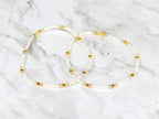 Two delicate beaded bracelets. Each bracelet features small white beads interspersed with gold-colored beads and pink beads. The bracelets are displayed on a white and gray marble surface.