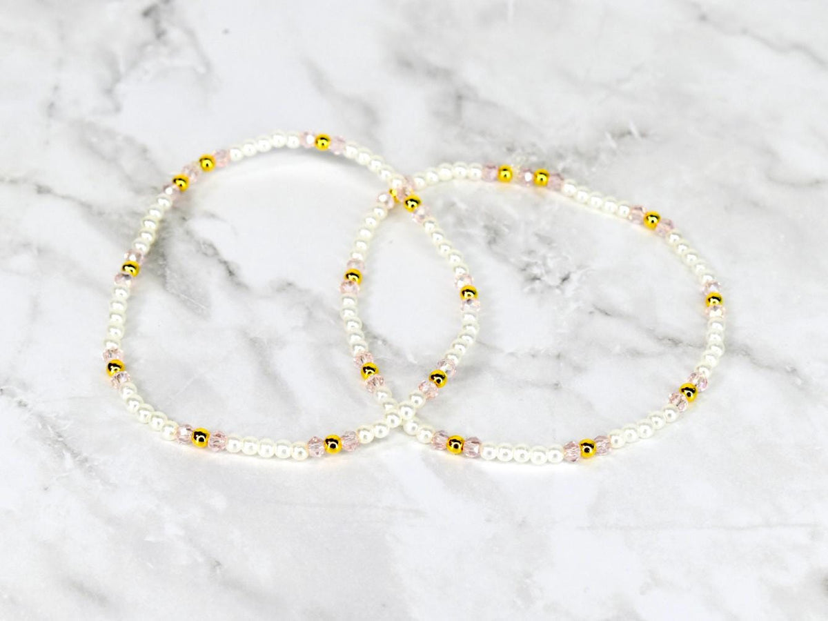 Two delicate beaded bracelets. Each bracelet features small white beads interspersed with gold-colored beads and pink beads. The bracelets are displayed on a white and gray marble surface.