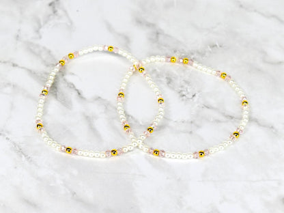 Two delicate beaded bracelets. Each bracelet features small white beads interspersed with gold-colored beads and pink beads. The bracelets are displayed on a white and gray marble surface.