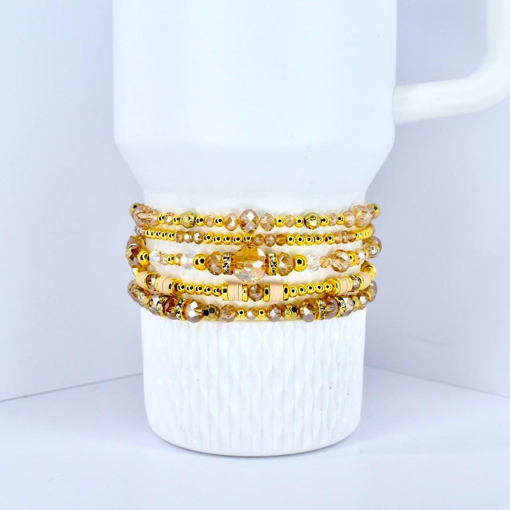 A stack of gold-toned beaded bracelets with various sizes and shapes of beads. The bracelets are displayed on a white mug with a textured surface. The beads have a mix of clear and amber colors.
