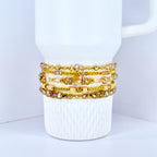 A stack of gold-toned beaded bracelets with various sizes and shapes of beads. The bracelets are displayed on a white mug with a textured surface. The beads have a mix of clear and amber colors.