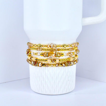 A stack of gold-toned beaded bracelets with various sizes and shapes of beads. The bracelets are displayed on a white mug with a textured surface. The beads have a mix of clear and amber colors.