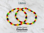 Two beaded bracelets with red, yellow, green, and black beads. The bracelets are labeled "Freedom" and are part of the Calathea Collective. The bracelets are shown on a white and gray marble surface.