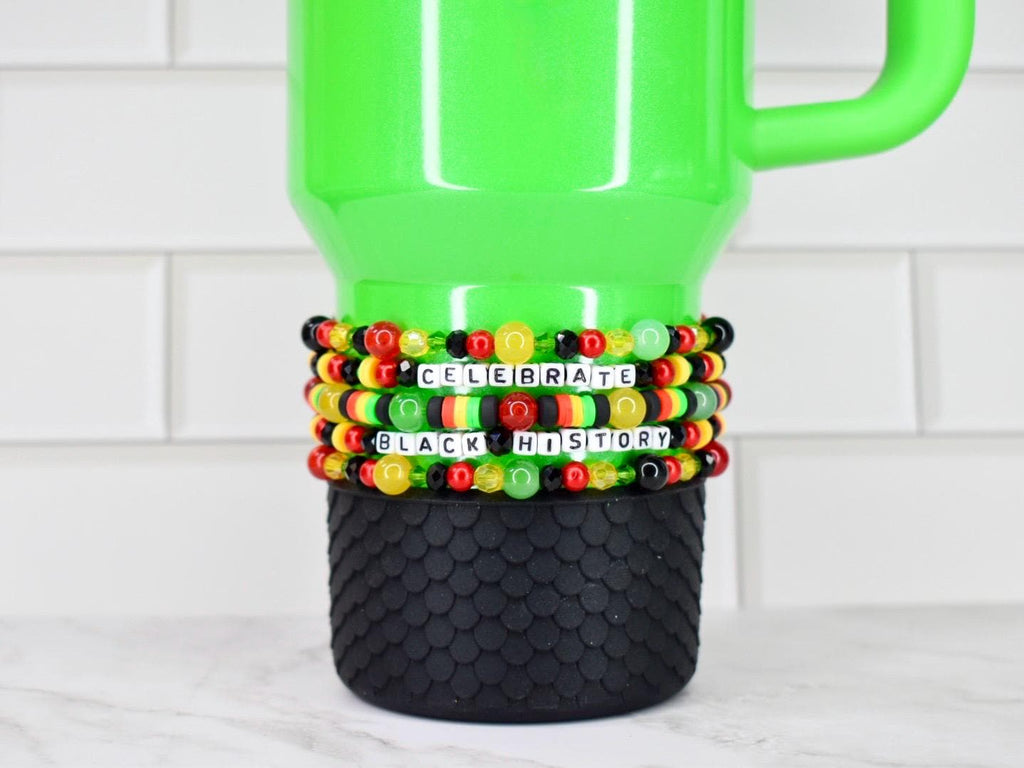 A green tumbler with a black textured base. The tumbler is adorned with Black History Month themed beaded bracelets in the colors of the Pan-African flag, spelling out "CELEBRATE BLACK HISTORY" in white letter beads. The background is a white tile.