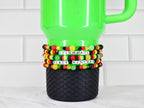 A green tumbler with a black textured base. The tumbler is adorned with Black History Month themed beaded bracelets in the colors of the Pan-African flag, spelling out "CELEBRATE BLACK HISTORY" in white letter beads. The background is a white tile.