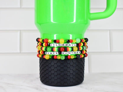 A green tumbler with a black textured base. The tumbler is adorned with Black History Month themed beaded bracelets in the colors of the Pan-African flag, spelling out "CELEBRATE BLACK HISTORY" in white letter beads. The background is a white tile.