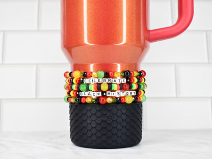 A red travel mug with a black textured base, adorned with beaded bracelets. The bracelets feature black, red, yellow, and green beads, spelling out "CELEBRATE BLACK HISTORY" in white letter beads.