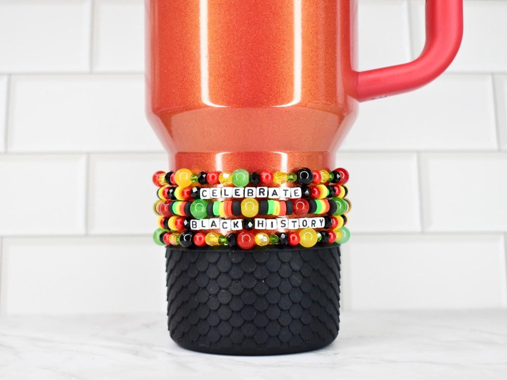 A red travel mug with a black textured base, adorned with beaded bracelets. The bracelets feature black, red, yellow, and green beads, spelling out "CELEBRATE BLACK HISTORY" in white letter beads.