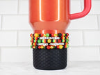 A red travel mug with a black textured base, adorned with beaded bracelets. The bracelets feature black, red, yellow, and green beads, spelling out "CELEBRATE BLACK HISTORY" in white letter beads.