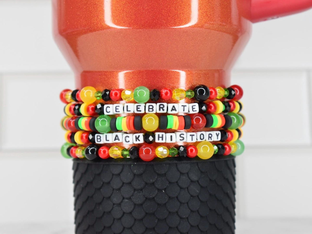 A stack of colorful beaded bracelets with the words "CELEBRATE BLACK HISTORY" spelled out in white letter beads. The bracelets are in red, yellow, green, and black. The bracelets are around a red and black tumbler.