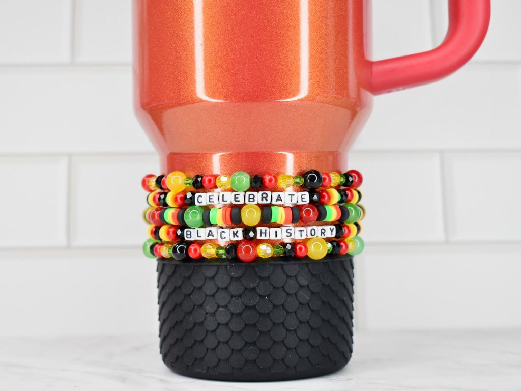 A red and orange tumbler with a black textured base. The tumbler is adorned with beaded bracelets in the colors of the Pan-African flag, spelling out "CELEBRATE BLACK HISTORY". The bracelets are stacked around the tumbler.