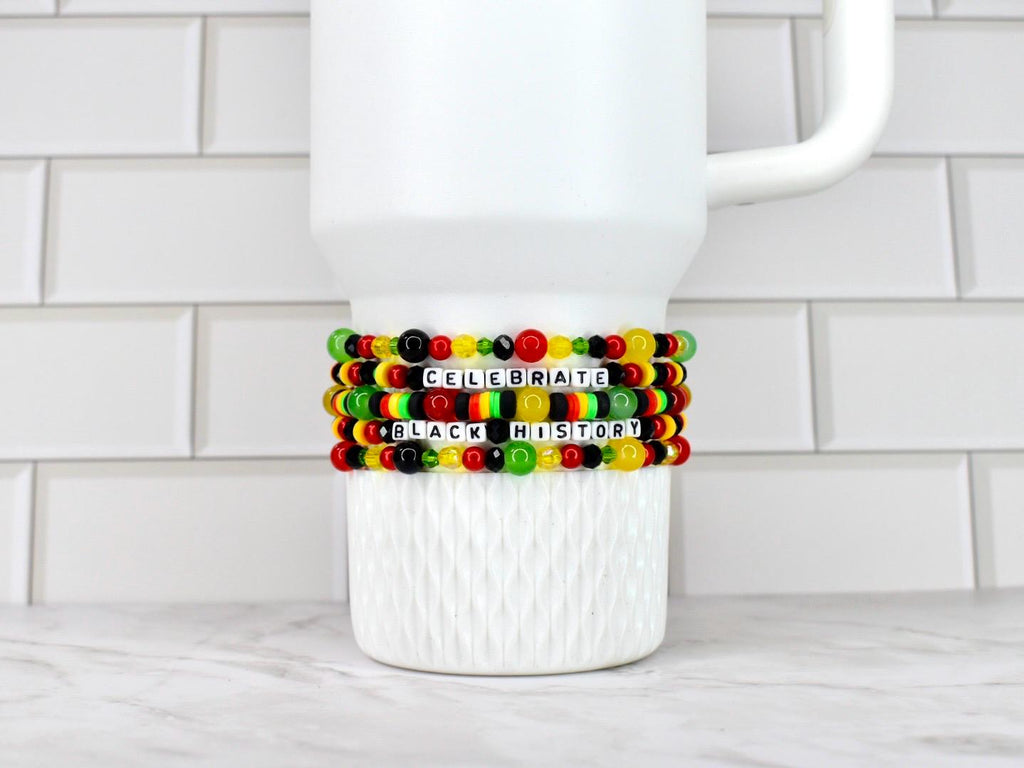 A white ceramic mug with three beaded bracelets wrapped around it. The bracelets are in the colors of the Pan-African flag, with the words "CELEBRATE BLACK HISTORY" spelled out in white letter beads.