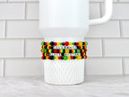 A white ceramic mug with three beaded bracelets wrapped around it. The bracelets are in the colors of the Pan-African flag, with the words "CELEBRATE BLACK HISTORY" spelled out in white letter beads.