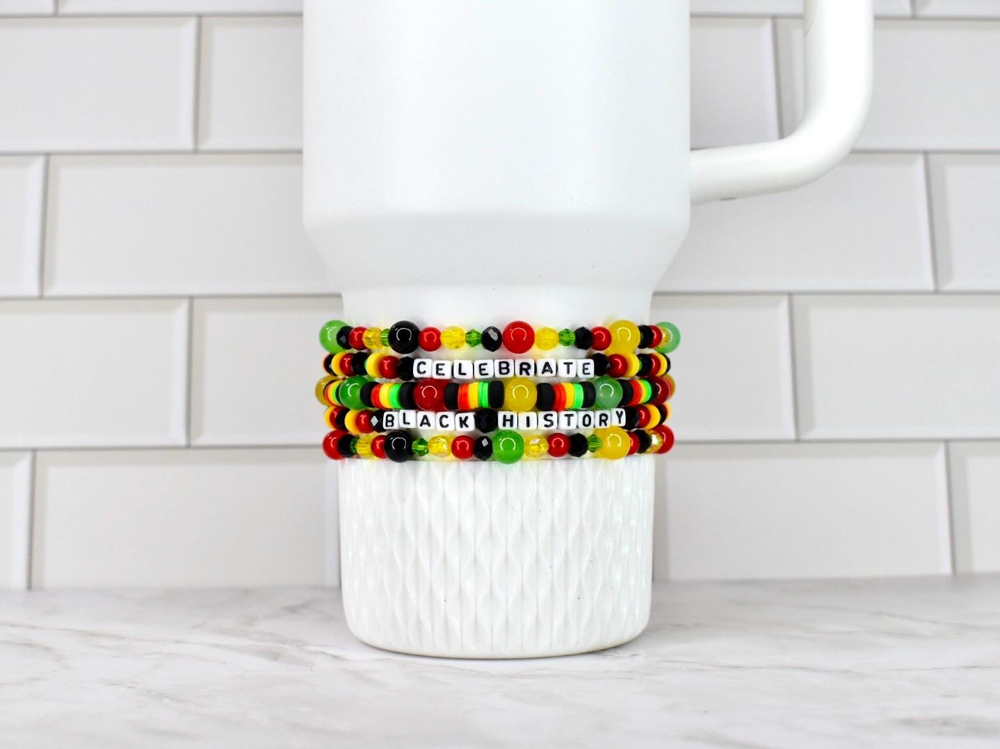 A white ceramic mug with three beaded bracelets wrapped around it. The bracelets are in the colors of the Pan-African flag, with the words "CELEBRATE BLACK HISTORY" spelled out in white letter beads.