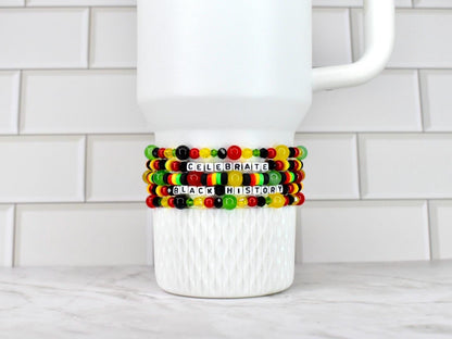 A white ceramic mug with three beaded bracelets wrapped around it. The bracelets are in the colors of the Pan-African flag, with the words "CELEBRATE BLACK HISTORY" spelled out in white letter beads.