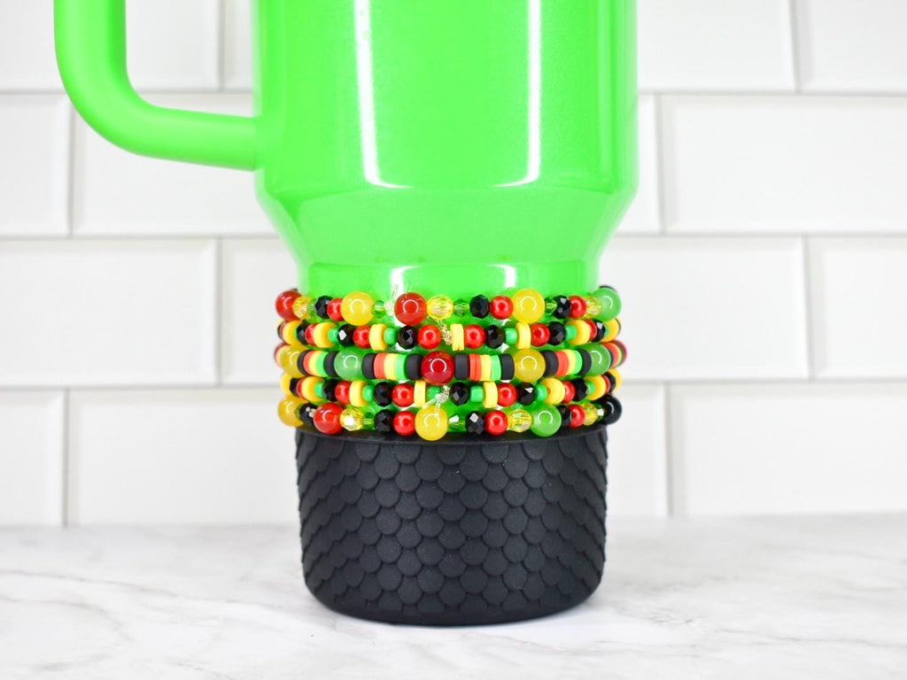 A bright green travel mug with a black textured base and a beaded sleeve. The sleeve features red, yellow, black, and green beads. The mug has a handle and is set against a white tile background.
