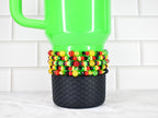 A bright green travel mug with a black textured base and a beaded sleeve. The sleeve features red, yellow, black, and green beads. The mug has a handle and is set against a white tile background.