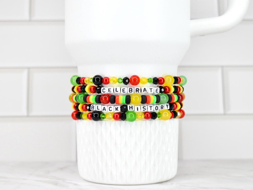 Four beaded bracelets with the words "CELEBRATE BLACK HISTORY" in white letter beads. The bracelets feature red, yellow, green, and black beads. The bracelets are stacked on a white ceramic mug.