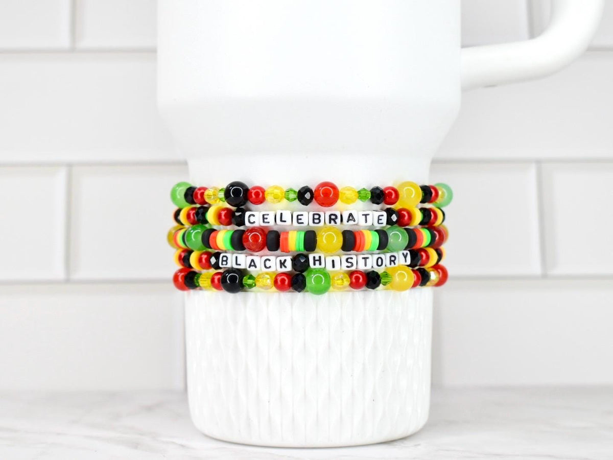 Four beaded bracelets with the words "CELEBRATE BLACK HISTORY" in white letter beads. The bracelets feature red, yellow, green, and black beads. The bracelets are stacked on a white ceramic mug.