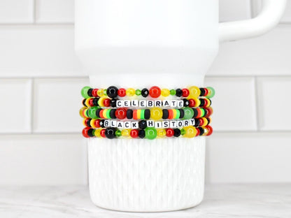 Four beaded bracelets with the words "CELEBRATE BLACK HISTORY" in white letter beads. The bracelets feature red, yellow, green, and black beads. The bracelets are stacked on a white ceramic mug.
