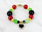 A beaded bracelet featuring red, green, black, and yellow beads. The bracelet has a gold-colored magnetic clasp and a heart-shaped charm with a black center and gold-colored frame. The beads are round and smooth.