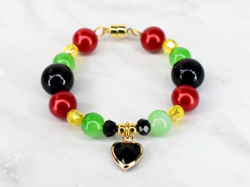 A beaded bracelet featuring red, green, black, and yellow beads. The bracelet has a gold-colored magnetic clasp and a heart-shaped charm with a black center and gold-colored frame. The beads are round and smooth.