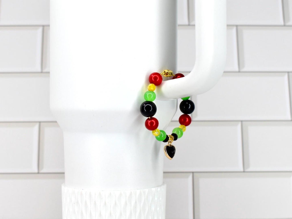 A beaded bracelet with red, yellow, green, and black beads. The bracelet has a gold-colored heart charm. The bracelet is displayed on a white mug handle against a white tile background.